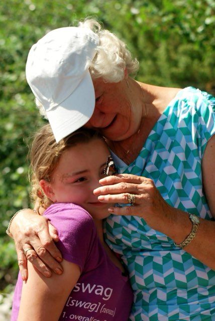 An older woman standing hugging a young girl, looking at a butterfly on the older woman's hand.